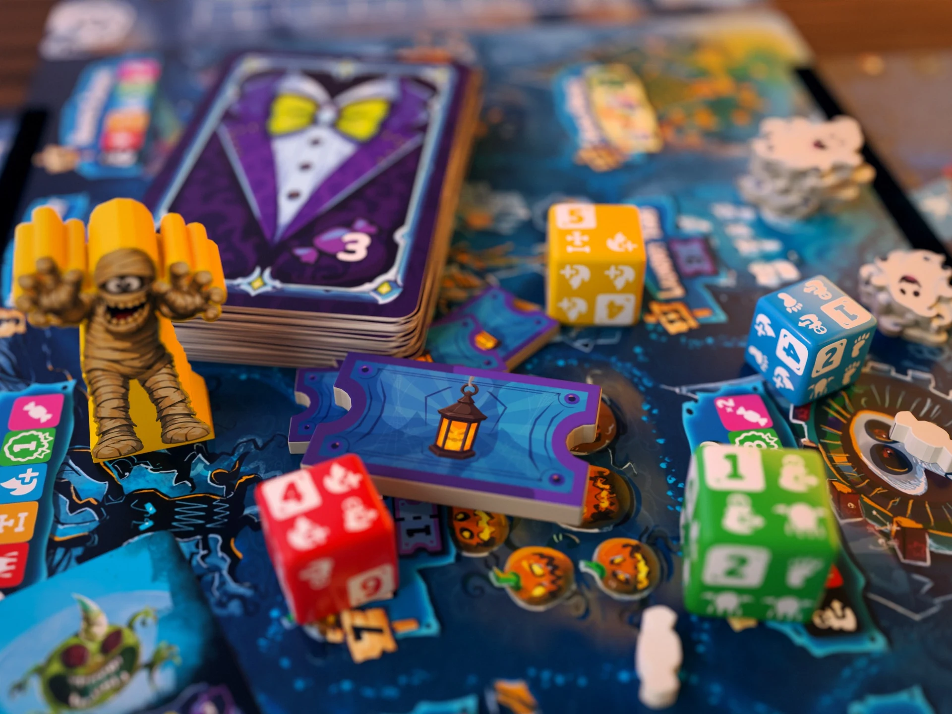 A yellow mummy Monster beside a deck of cards, lantern ticket tokens, and colorful Guest Dice on a player board